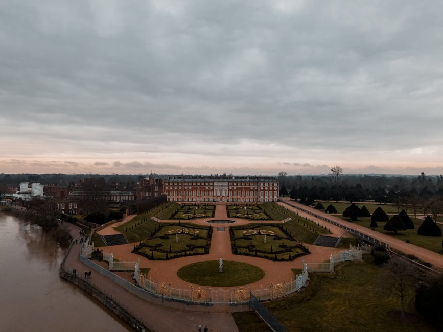 An aerial view of a stately garden with symmetrical flowerbeds bordered by manicured hedges, leading towards a grand historic building in the distance. The garden features a central circular lawn with