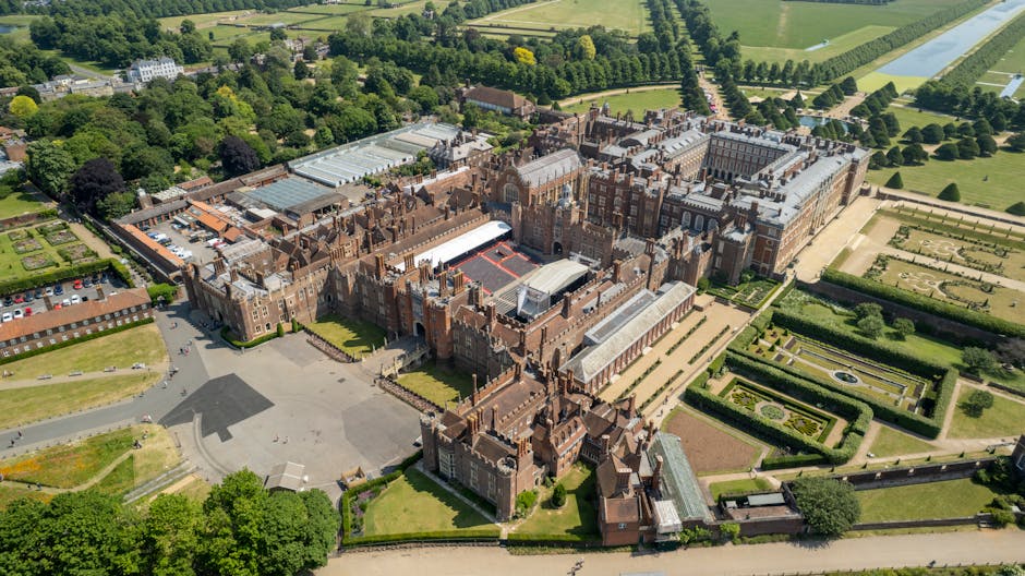 An aerial view of a stately garden with symmetrical flowerbeds bordered by manicured hedges, leading towards a grand historic building in the distance. The garden features a central circular lawn with