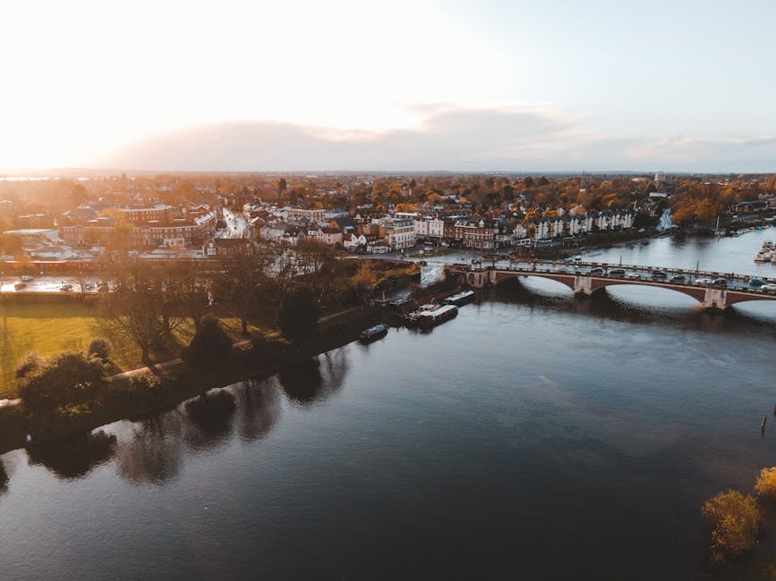 An aerial view of the Thames River flowing through Kingston with a stone bridge crossing the water. On the riverbanks, there are lush green lawns and trees with textured leaves, some partially in shad