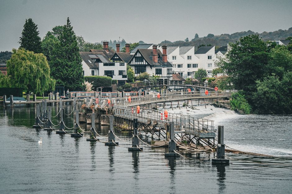 An aerial view of the Thames River flowing through Kingston with a stone bridge crossing the water. On the riverbanks, there are lush green lawns and trees with textured leaves, some partially in shad
