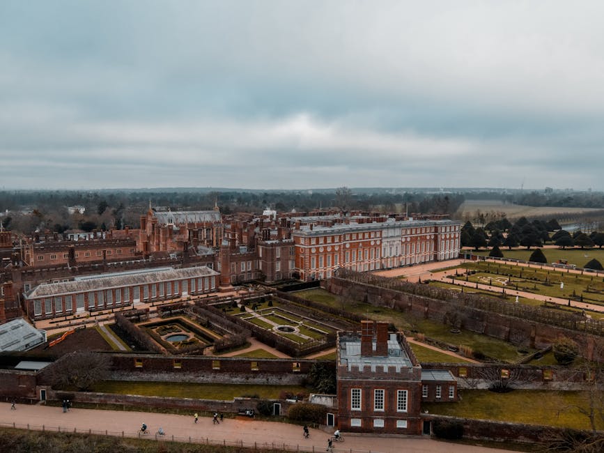 An expansive view of a historic landscaped garden with neatly maintained lawns, geometric flower beds, and trimmed hedges, bordered by a pathway with pedestrians. In the background, there is a large, 