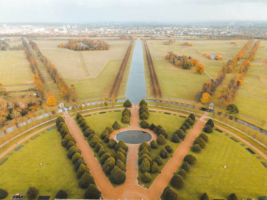 An expansive view of a historic landscaped garden with neatly maintained lawns, geometric flower beds, and trimmed hedges, bordered by a pathway with pedestrians. In the background, there is a large, 